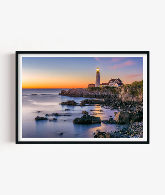 Bring peaceful coastal atmosphere to your space with this rocky lighthouse print, featuring a glowing beacon, rugged shoreline, calm ocean water and a soft orange sunset sky for bathrooms, bedrooms, living rooms, offices, beach houses and gallery walls. 